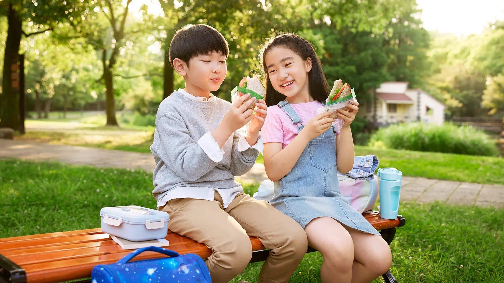 Two school age children sitting on a bench outside eating sandwiches from Ziploc® bags